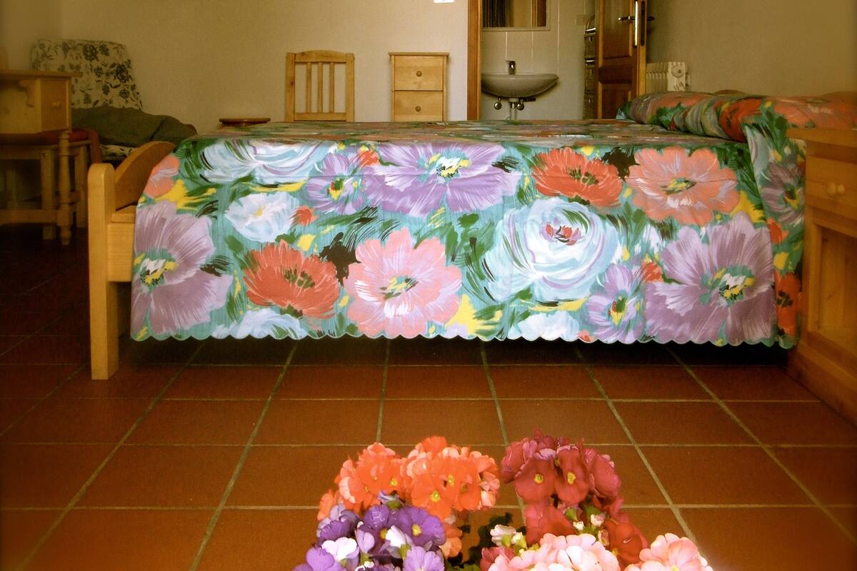 Une chambre avec une couvre-lit à motifs floraux, une chaise en bois, une table de chevet et un petit vase de fleurs posé sur le sol.