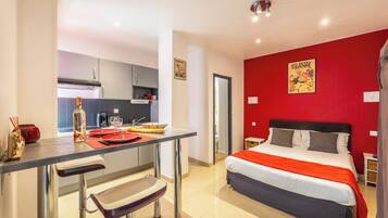 Habitación de hotel moderna con zona de cocina, cama con colcha roja y blanca, y pared de acento en color rojo.