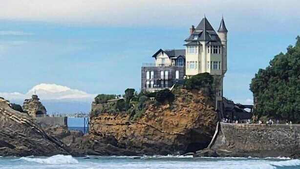 Un bâtiment situé sur la côte, doté d’une tour, entouré de vagues et de surfeurs.