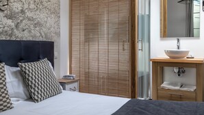 Une chambre à coucher comprenant un lit, une table de chevet, un store en bois, une salle de bain avec un lavabo et un miroir, et un mur orné d’un motif texturé.