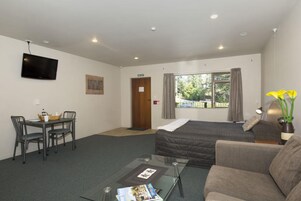 Quarto de hotel com cama, sofá, mesa de vidro, televisão, janela com cortinas e porta.