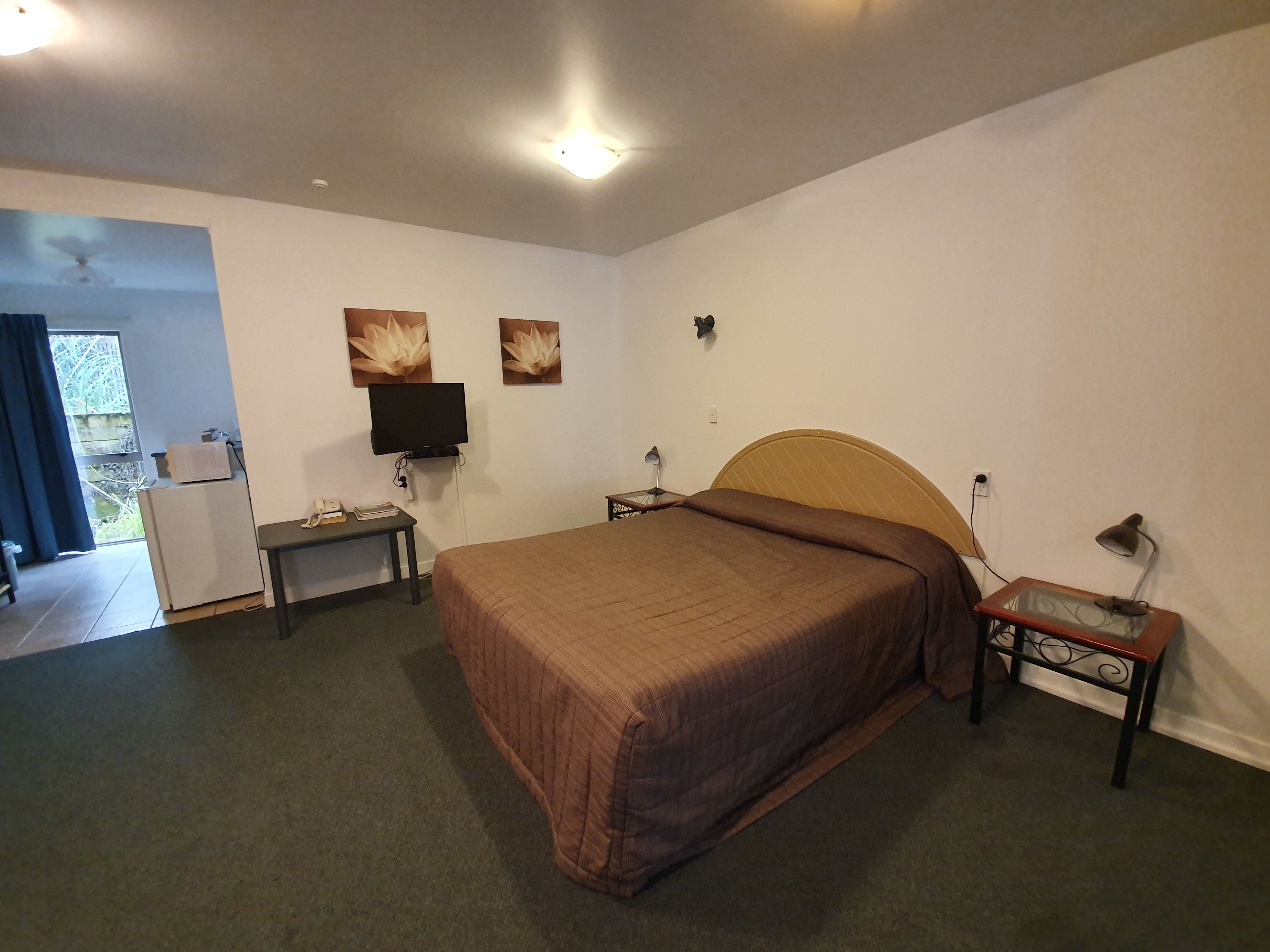 A bedroom with a large bed, a desk with a computer, a TV, and a small table with a lamp.