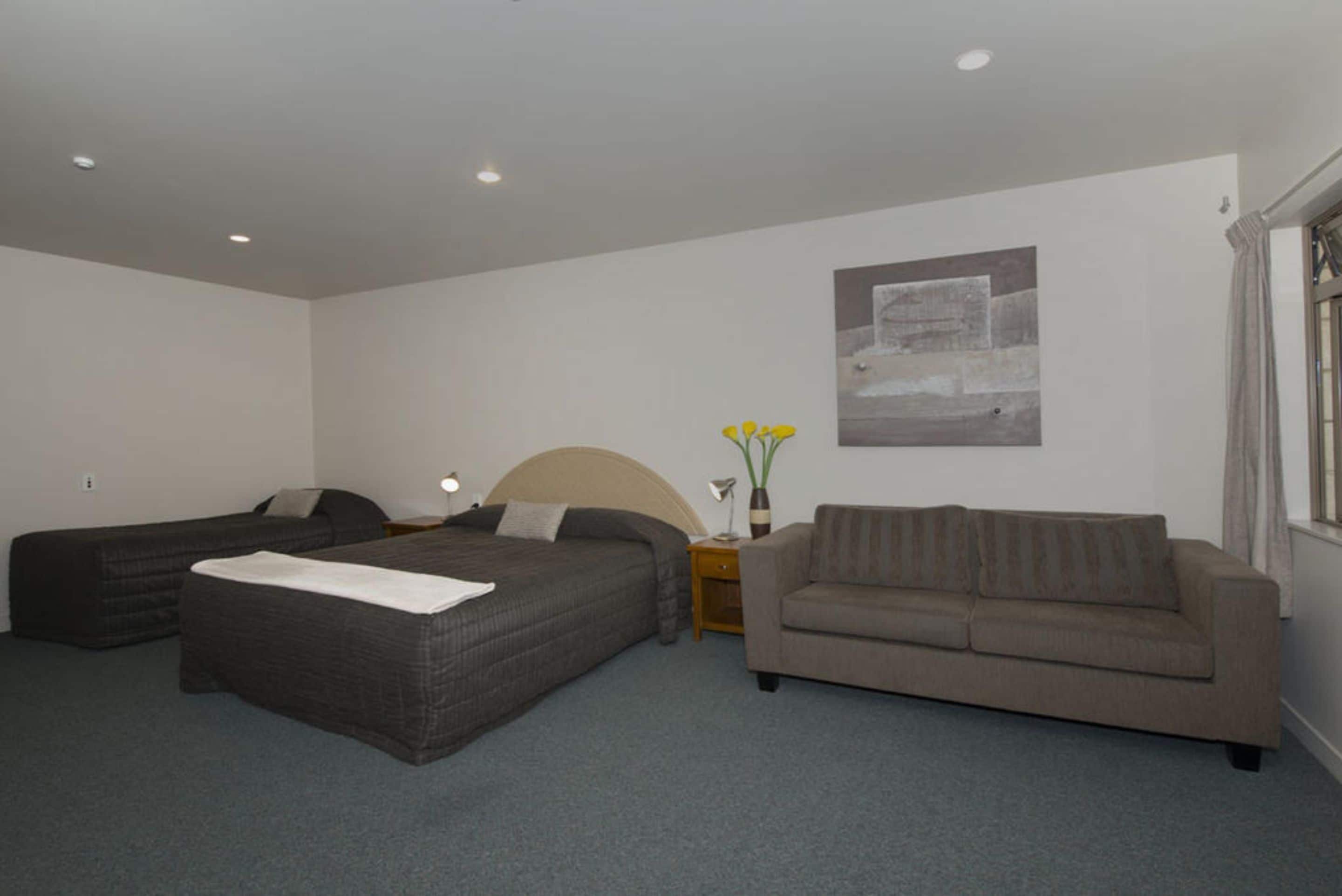 A hotel room with two single beds, a sofa, a nightstand with a lamp, and a vase with flowers.