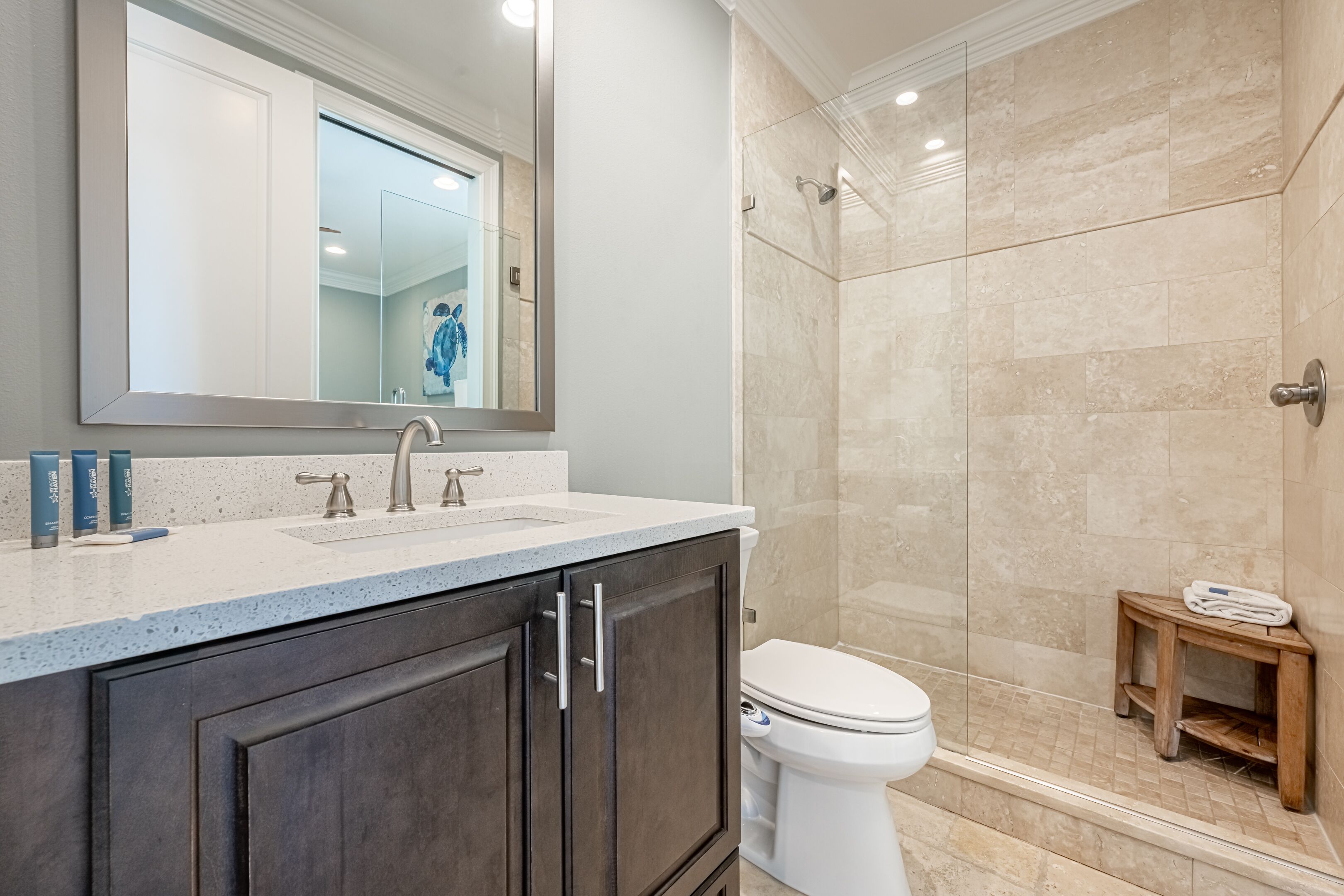 A modern bathroom with a large mirror, a sink, and a toilet. There is a shower area with a glass door and a small wooden stool.