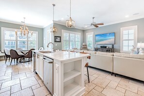 A modern kitchen with a central island, dining area with chairs, a sofa, and a TV mounted on the wall. - Two Golf Carts ~ Resort Beach ~ Community Pool (Miramar Beach)