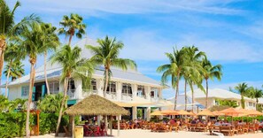 Un hotel frente a la playa con un edificio grande, palmeras y zonas de estar al aire libre.