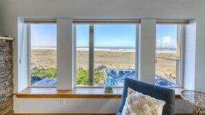 Ein Zimmer mit großen Fenstern mit Blick auf einen Strand, einer Steinwand und einem Korbsessel mit Kissen.