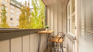 Een balkon met een kleine houten tafel en twee stoelen, een plant in pot en uitzicht op gebouwen en bomen.