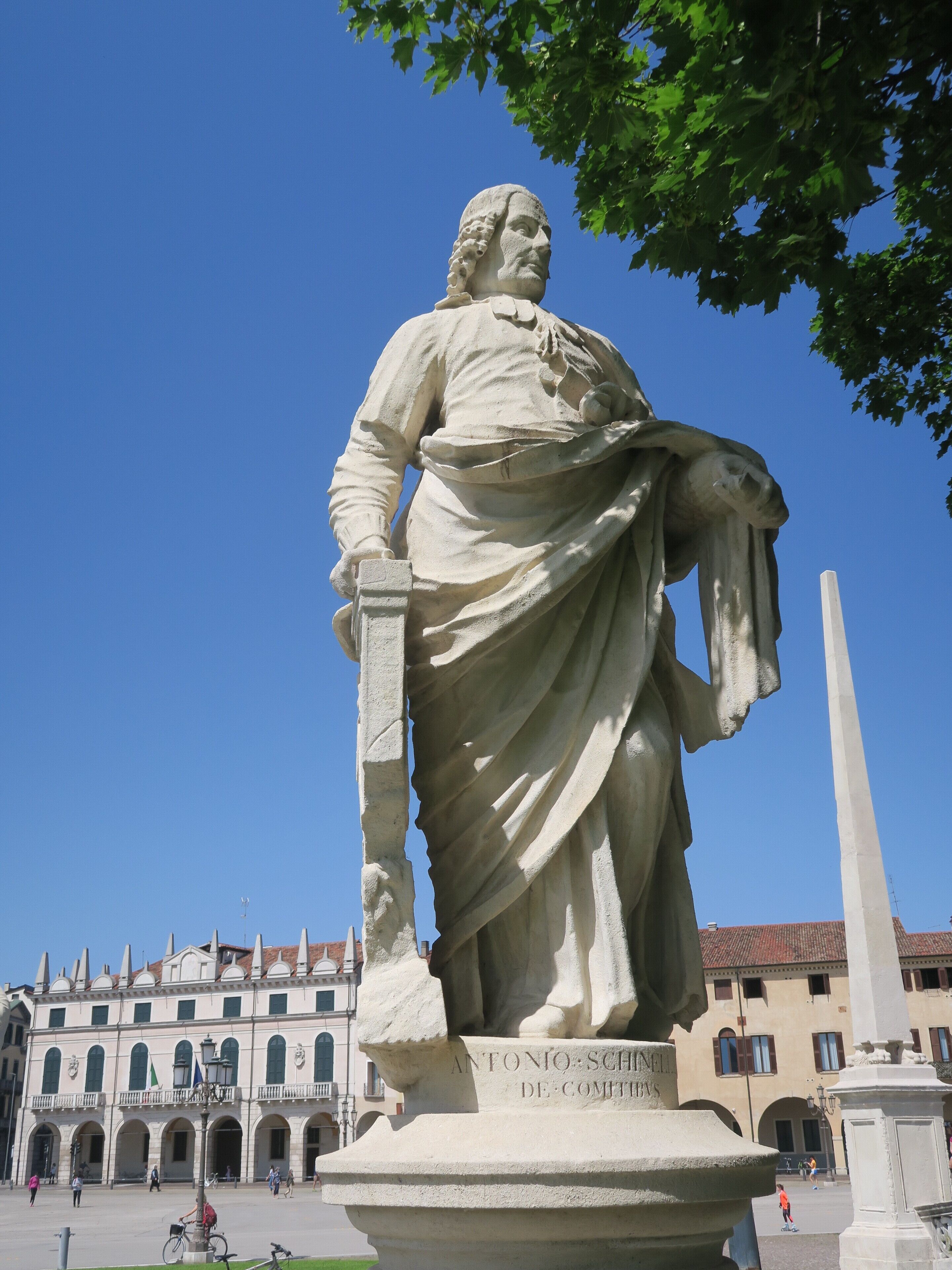 En statue af en historisk person i en offentlig plads med en bygning i baggrunden.
