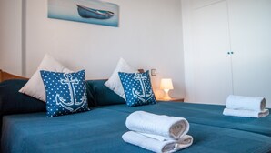 A neatly made bed with blue bedding and white pillows, a bedside lamp, and a painting of a boat on the wall. - Apartment Aldeas Taray Club 4/5 Admer (La Manga del Mar Menor)