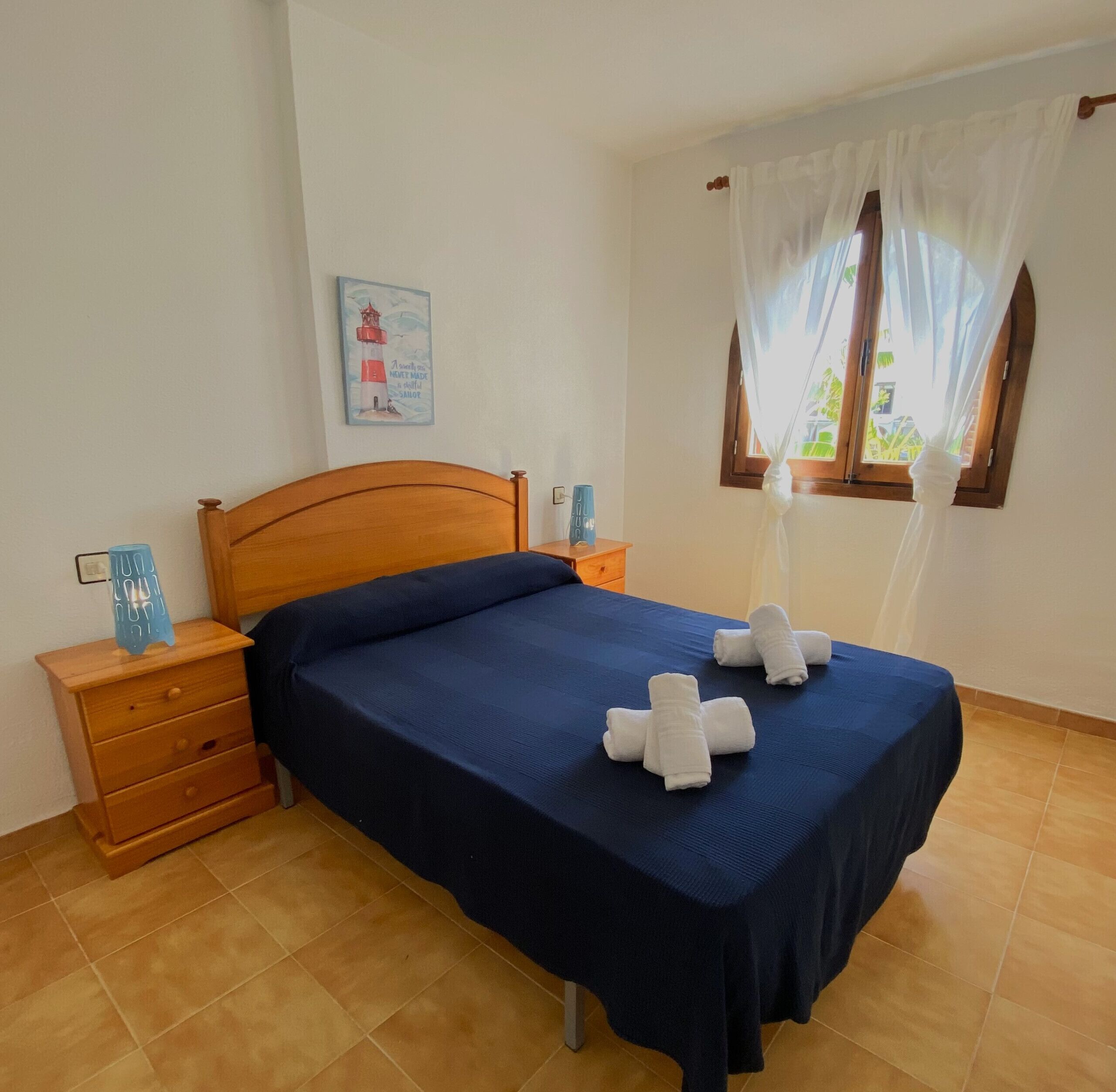 Una cama bien hecha con colcha azul y toallas blancas, cabecero de madera, mesita de noche con lámpara y ventana con cortinas translúcidas.