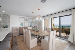 Un coin repas avec une table en bois et des chaises rembourrées, un lustre et une vue sur l’océan grâce à de grandes portes coulissantes.