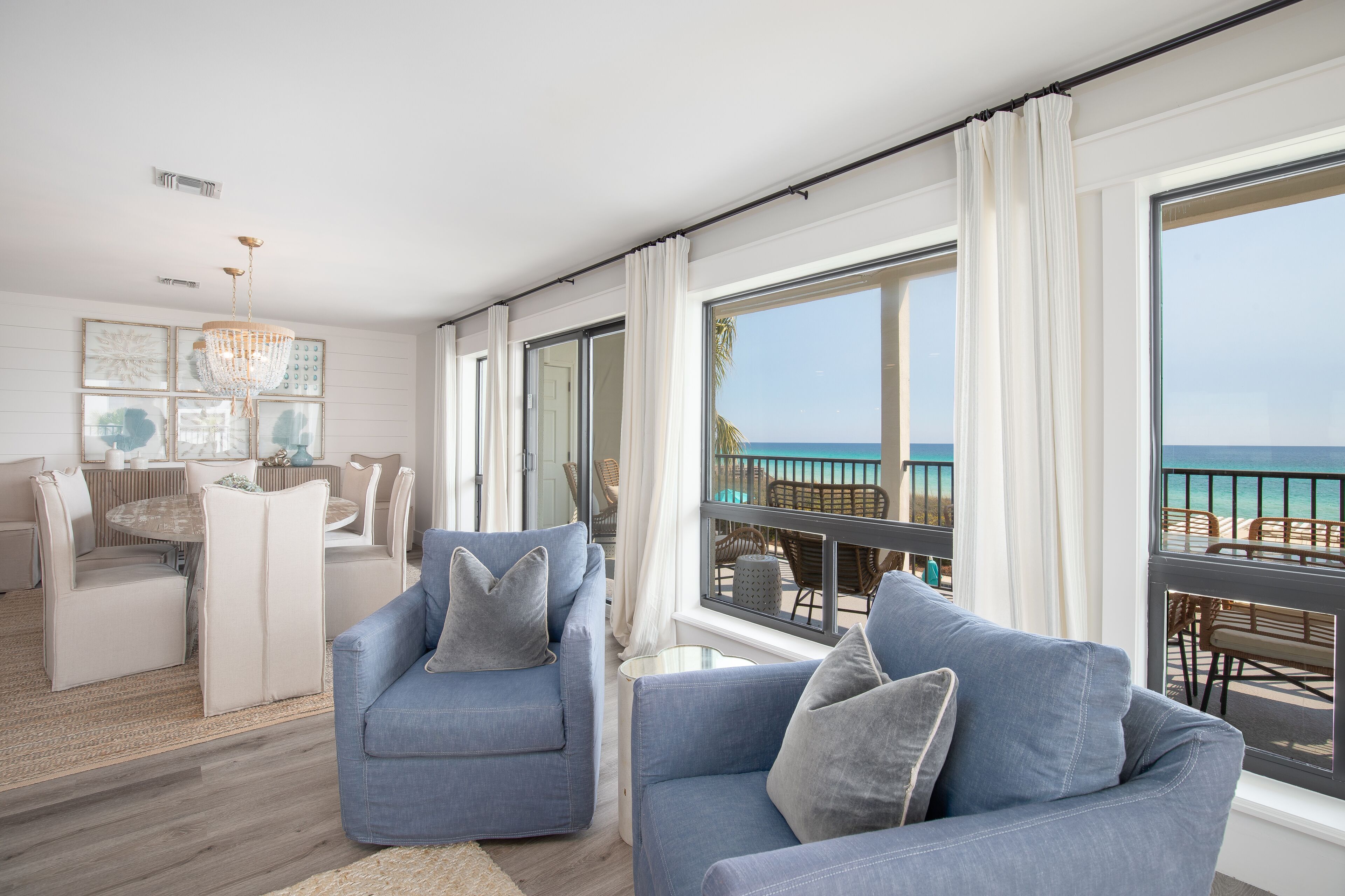 Ein modernes Wohnzimmer mit großem Fenster mit Blick auf einen Strand, ein Essbereich mit rundem Tisch und einer Chandelier.