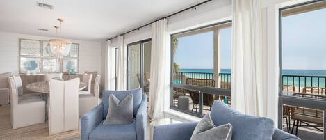 Ein modernes Wohnzimmer mit großem Fenster mit Blick auf einen Strand, ein Essbereich mit rundem Tisch und einer Chandelier.