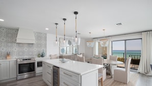 Una cocina moderna con armarios blancos, electrodomésticos de acero inoxidable y una isla central. Hay un comedor con mesa y sillas, y un balcón con vista al mar.