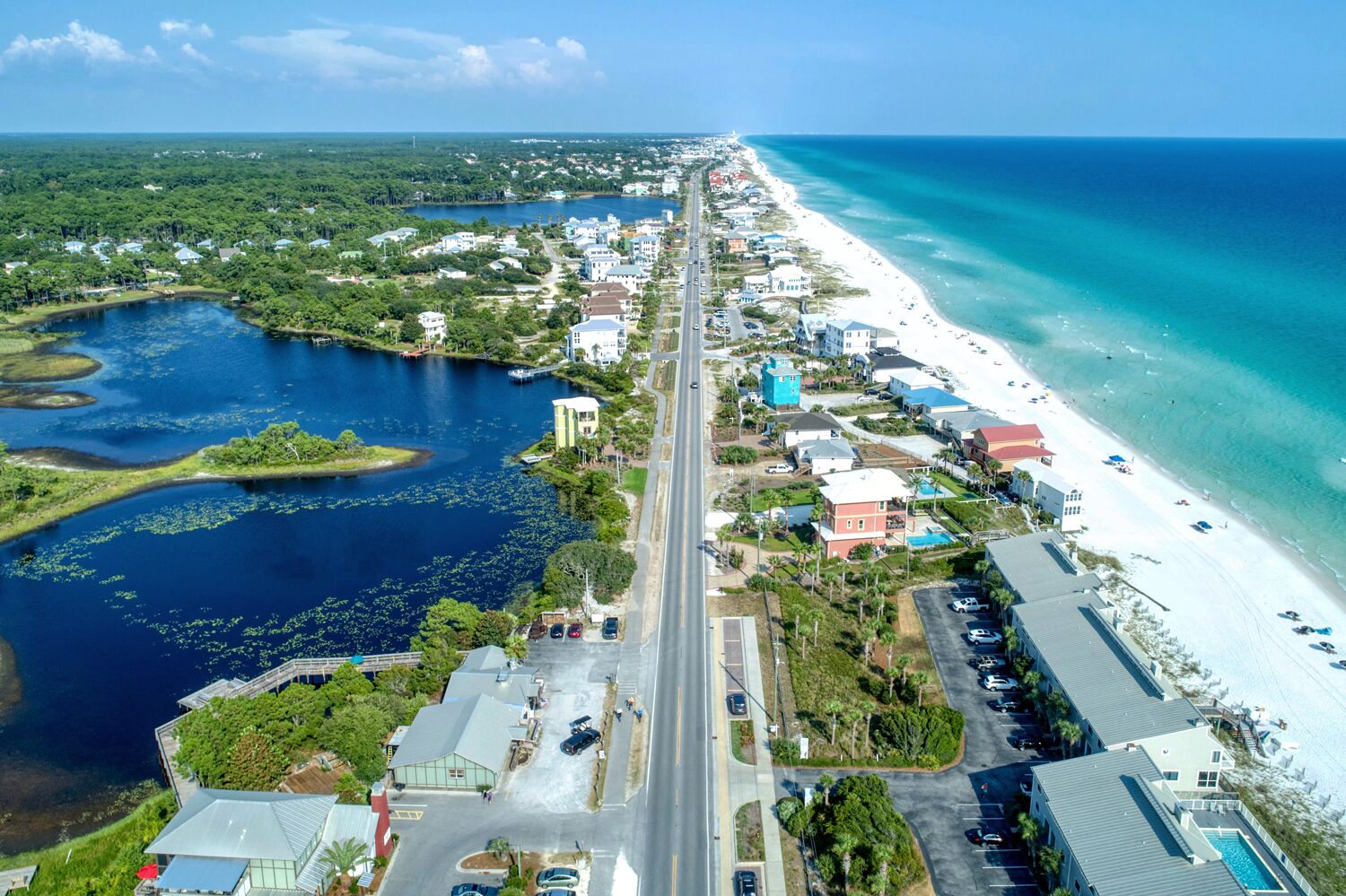 An aerial view of a coastal town with a beach, a marina, and a road running through the center.
