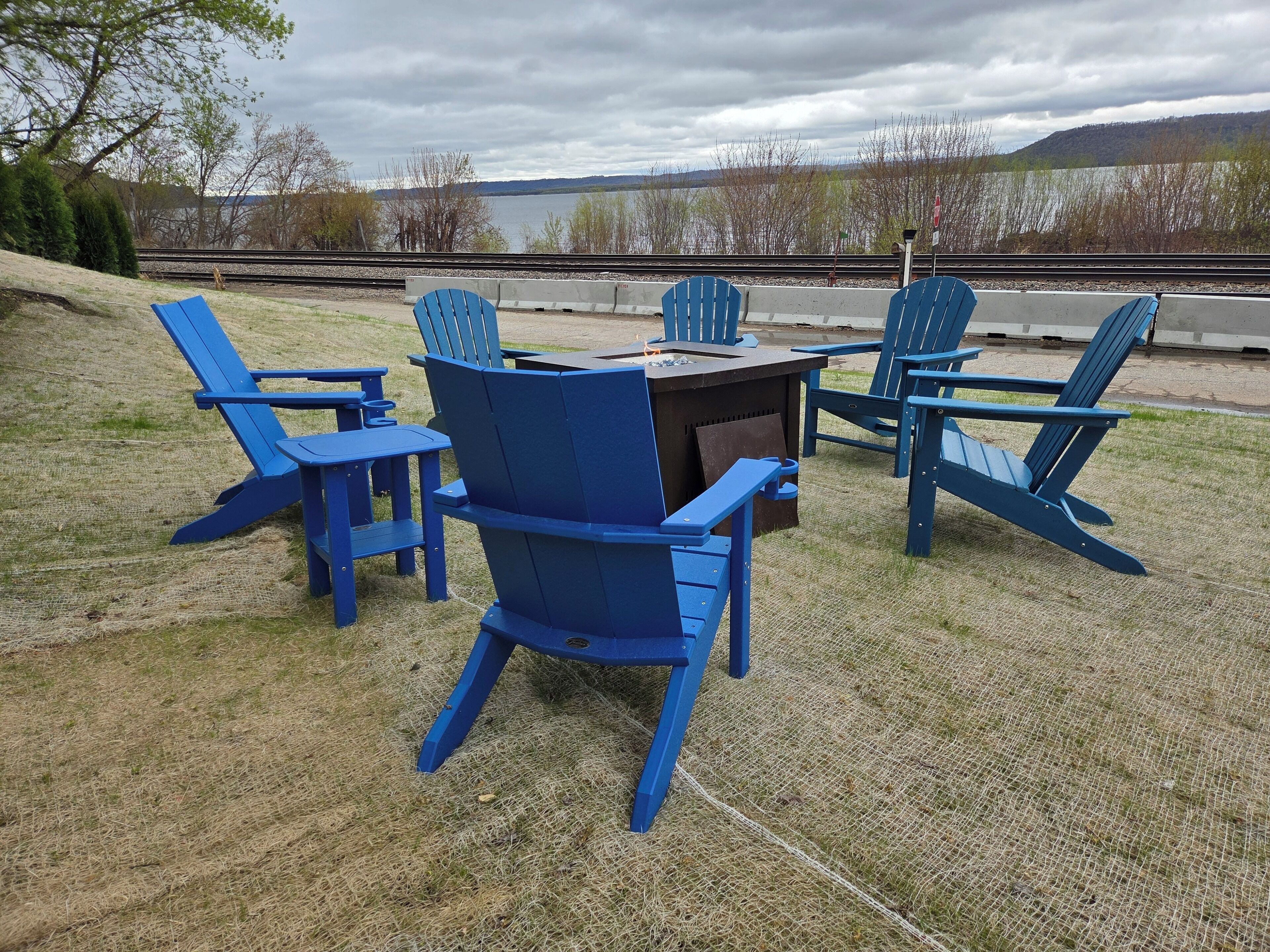 Un'area picnic con poltrone Adirondack blu e un tavolo, con vista su un lago e dei binari ferroviari.