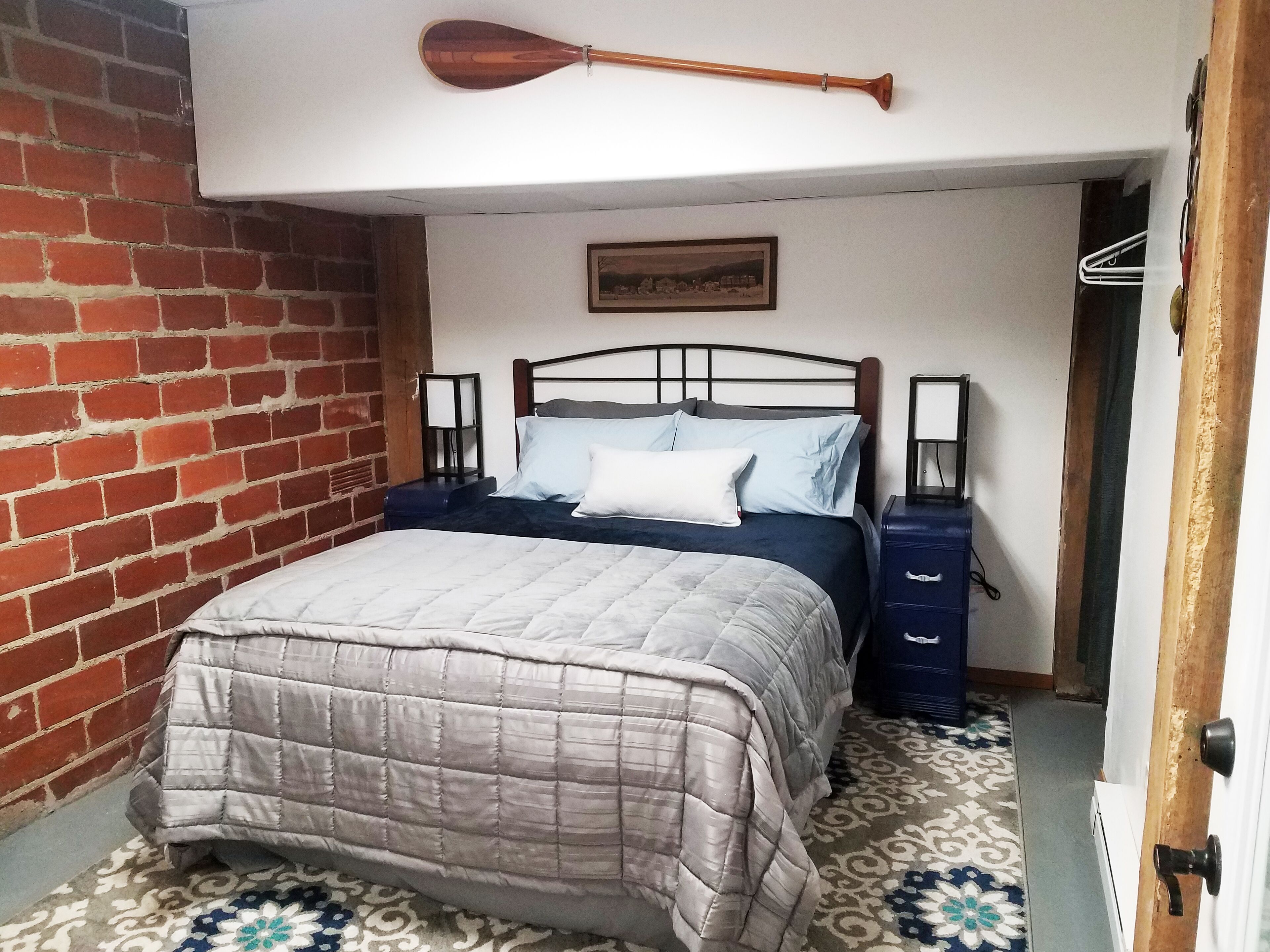 Una habitación con una cama, una pala de madera, un muro de ladrillo, un suelo estampado y una puerta.