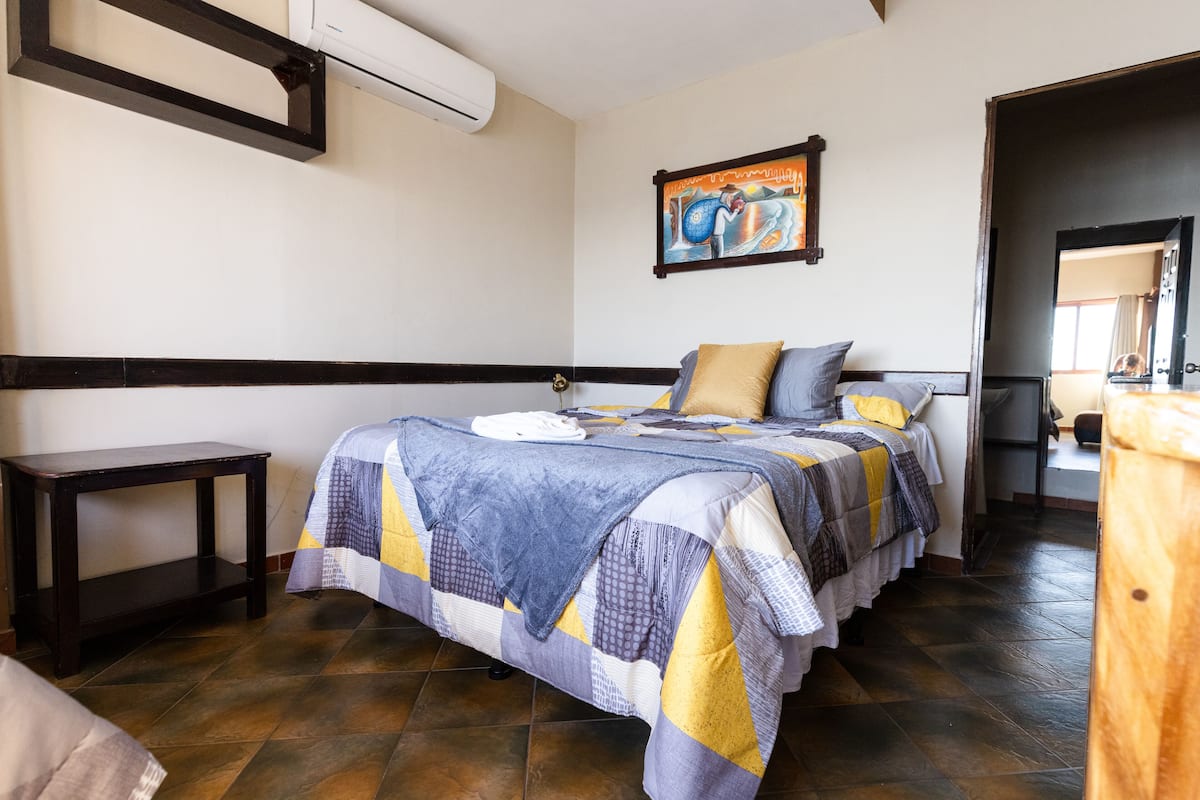 A bedroom with a bed, a bedside table, a wall-mounted air conditioner, and a painting on the wall.