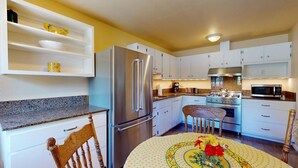 A compact kitchen with white cabinets, a stainless steel refrigerator, a microwave, and a stove. There is a dining table with a floral tablecloth and a chair.