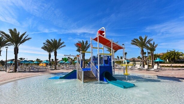 Area giochi d'acqua con scivoli e struttura per arrampicata, circondata da palme e sdraio.
