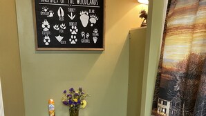 A bathroom with a toilet, a trash can, a toilet paper holder, a framed sign with animal paw prints, and a curtain with a forest scene. - Attractive Country Get-a (Abbeville)