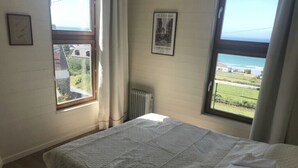 A bedroom with a bed, a radiator, a window with a view of the sea, and a framed picture on the wall.