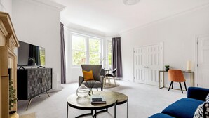 Una sala moderna con sofá, sillón y mesa de centro. Hay un televisor sobre un mueble bajo, una chimenea y una ventana con cortinas.