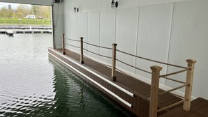 A swimming pool with wooden deck and rope barriers, surrounded by white walls and a view of a dock and greenery. - Waterfront Villa with Boathouse in Alexandria Bay (Alexandria Bay)
