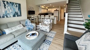 A modern living room with a grey sofa, a coffee table with a tray, and a patterned rug. A staircase leads to a kitchen area with white cabinets and a microwave. - Waterfront Villa with Boathouse in Alexandria Bay (Alexandria Bay)