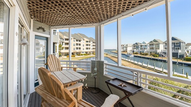 A covered outdoor area with a wooden table and chairs, a small table with a tray, and a view of a canal with buildings in the background. - Dock Holiday: Canalfront condo, Water Views, Fisherman's dream with Boat Dock (Manteo)