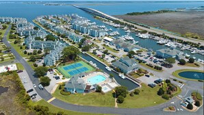 An aerial view of a coastal residential area with multiple apartment buildings, a marina with boats, and a large swimming pool. - Dock Holiday: Canalfront condo, Water Views, Fisherman's dream with Boat Dock (Manteo)
