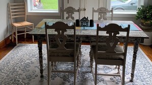Ein Essbereich mit Tisch, Stühlen und Teppich. Zwei Fenster bieten Blick auf eine grüne Landschaft und ein parkendes Auto. Über dem Tisch ist ein Deckenventilator zu sehen.
