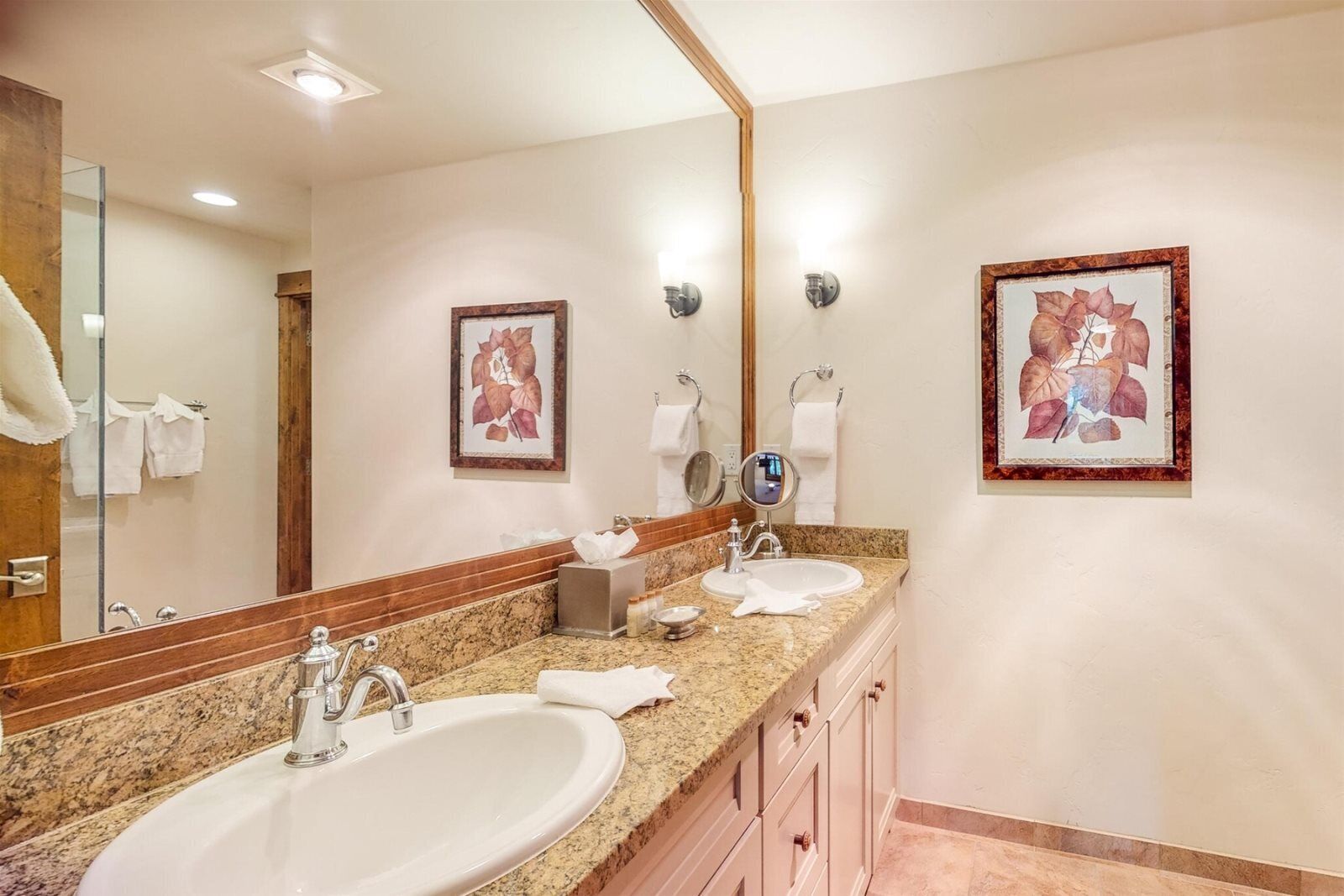 A hotel bathroom with two sinks, a large mirror, and framed botanical artwork on the walls.