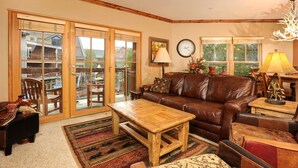 A cozy living room with a leather sofa, wooden coffee table, and a rug. There are two windows with curtains, a clock on the wall, and a chandelier.