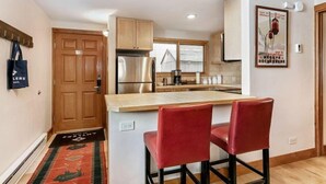 A compact kitchen with wooden cabinets, a stainless steel refrigerator, and a small island with two red bar stools. - 1 Bedroom Antlers Vacation Rental With Incredible Slopeside Views and Just a Short Walk to Gondola and Lionshead Village (Vail)