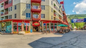 Un immeuble à plusieurs étages abritant un restaurant de pizzas au rez-de-chaussée, un distributeur automatique bien visible et ayant une chaîne de montagnes en toile de fond.