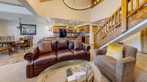 Un vaste espace de vie comprenant un canapé en cuir marron, une table basse en verre et un fauteuil gris. On aperçoit, à l’arrière-plan, un escalier en bois doté d’une rampe.