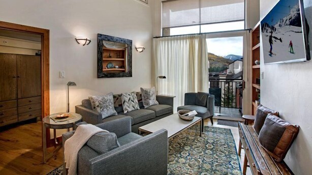 A spacious living room with a grey sofa, a coffee table, and a patterned rug. There is a wooden cabinet and a painting on the wall.