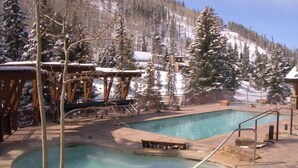 A pool area with a wooden deck, lounge chairs, and a view of snow-covered trees and mountains.
