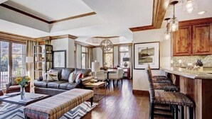 A spacious living room with a large sofa, a coffee table, and a dining area. There is a kitchen with wooden cabinets and a bar area. - Vail Ritz-carlton 3 Bedroom Residence in Lionshead Village (Vail)