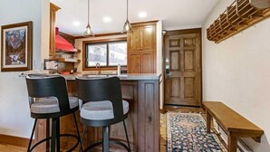 A compact kitchen with wooden cabinets, a central island with bar stools, pendant lighting, and a patterned rug. - 2 Bedroom Antlers Vacation Rental With Incredible Slopeside Views and Just a Short Walk to Gondola and Lionshead Village (Vail)