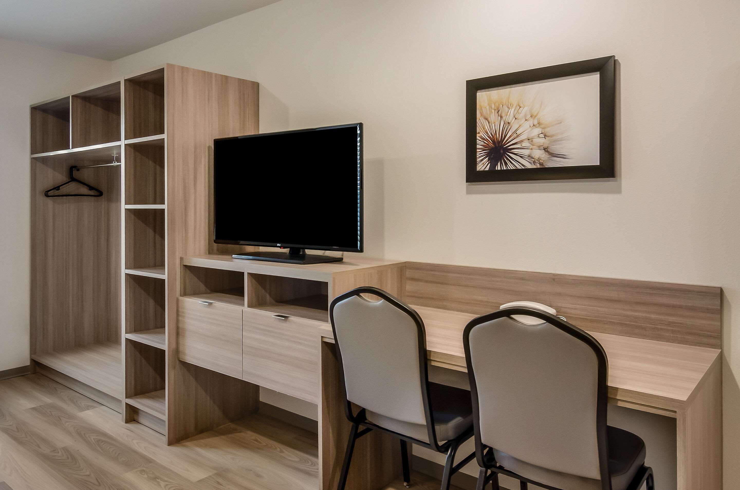 Una habitación moderna con un escritorio de madera, dos sillas, un televisor y un armario con varias repisas.