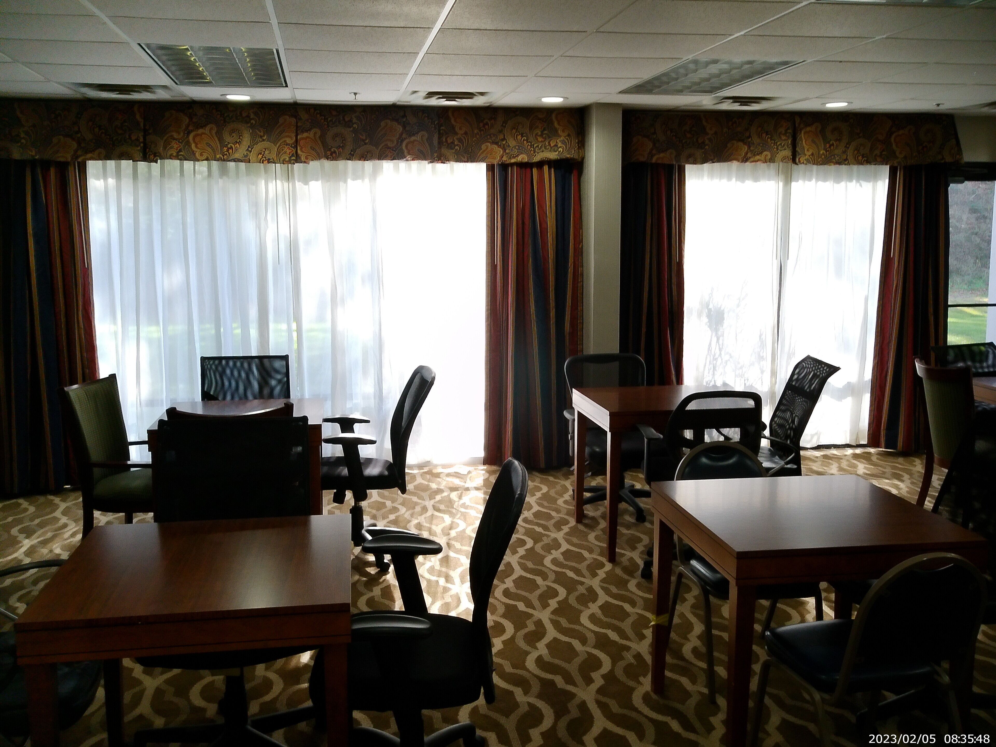 Habitación de hotel con zona de comedor que incluye una mesa y sillas, ventanas amplias con cortinas y alfombra estampada.