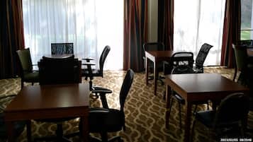 Habitación de hotel con zona de comedor que incluye una mesa y sillas, ventanas amplias con cortinas y alfombra estampada.