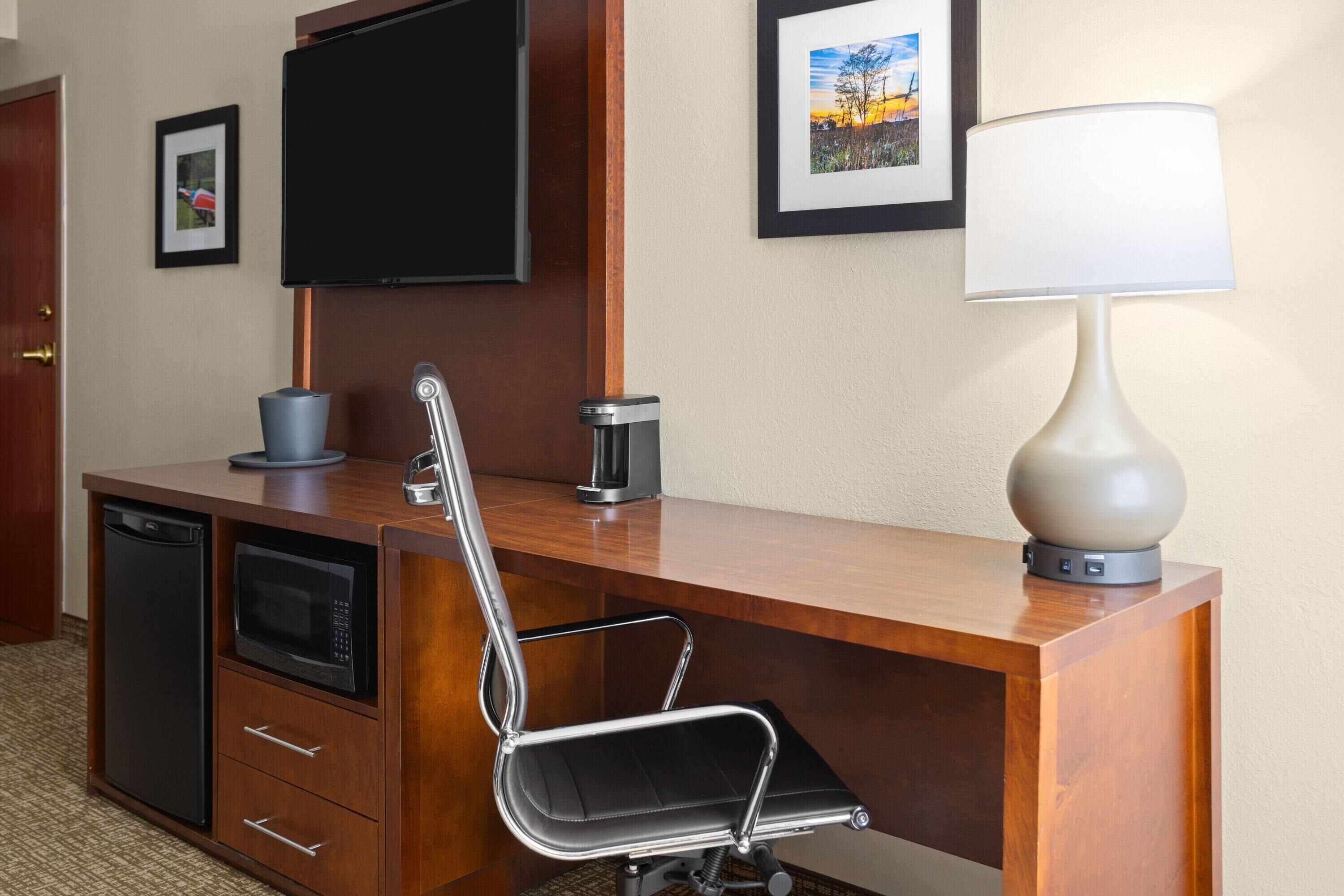 Habitación de hotel con un escritorio de madera, una silla de oficina negra, un televisor montado en la pared, una cafetera y una lámpara.