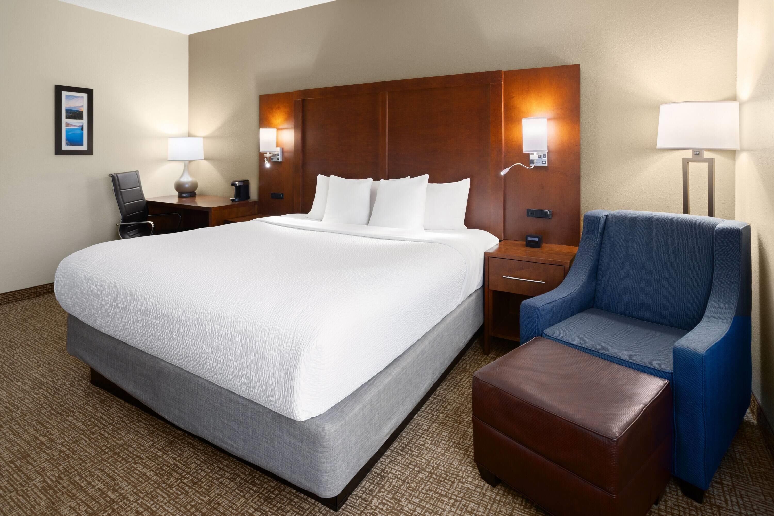 A hotel room with a large bed, a desk with a chair, a blue armchair, a brown ottoman, and two wall-mounted lamps.