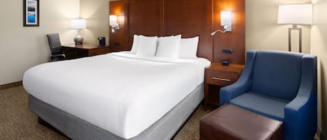 A hotel room with a large bed, a desk with a chair, a blue armchair, a brown ottoman, and two wall-mounted lamps.