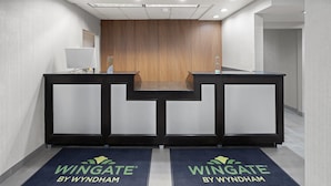 Uma área de recepção com dois balcões, uma parede de ripas de madeira e dois tapetes azuis com a marcação 'Wingate by Wyndham'.