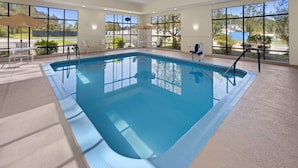Piscina interior com paredes de vidro transparente, chão de azulejos e alguns cadeiras de descanso e guarda-sóis.